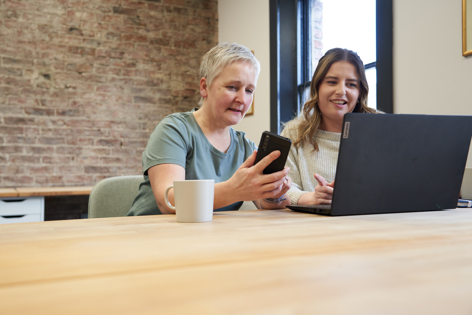 Alison O'Neill and Marianne Carey pictured sitting at a table speaking. Alison is holding a phone and Marianne is looking at a laptop.